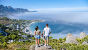 Couple standing on a rocky overlook, holding hands, gazing at a coastal town and blue sea under a clear sky with mountains in the distance.