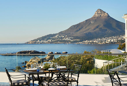 5 days Day 5 – Relax Before Departure Seaside outdoor dining terrace with a table set for four, overlooking the rocky shoreline and a distant mountain over the bay.