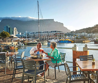 5days - Day 1 – Discover Cape Town An older couple dining at an outdoor waterfront cafe with boats and a marina in the background on a sunny day.