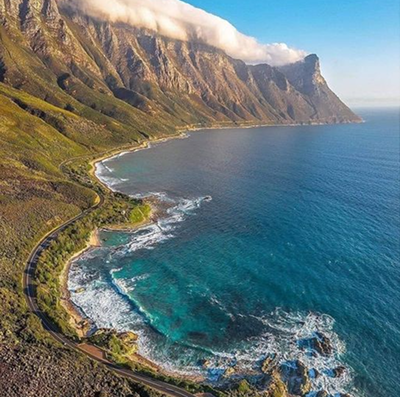 Winding coastal road along green cliffs, with turquoise ocean and white surf along rocky shore under a clear sky.