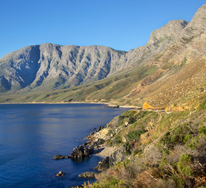 Clarence Drive – One of South Africa’s Hidden Gems Coastal landscape with blue sea on the left, rugged mountains in the background, and a dirt trail along the shore with a small orange tent on the hillside.