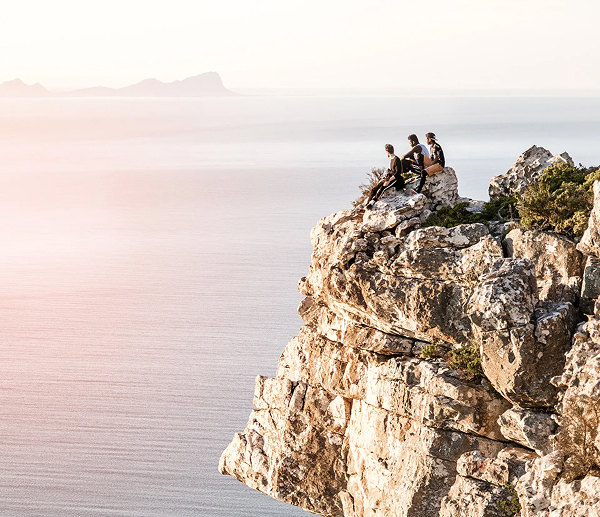 Packing for Winter Three hikers sit on a rocky cliff edge overlooking the calm sea at sunset.