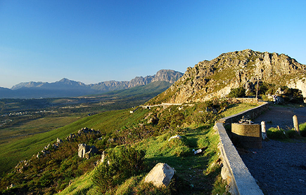 Sir Lowry’s Pass Viewpoint Sunlit mountain trail along a rocky hillside with a stone wall, sweeping valley and distant blue mountains under a clear sky.