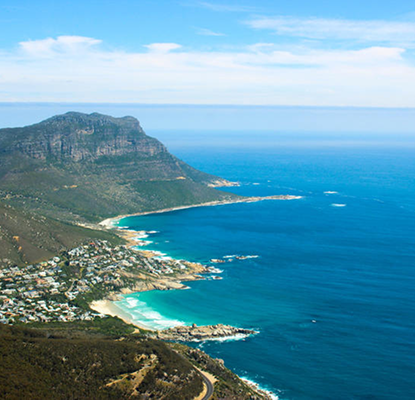The Cape Peninsula Scenic Route Aerial view of a coastal cliff, turquoise water, and a small town along a curved beach.