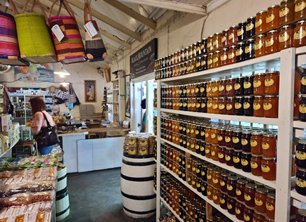 The Farm Stalls Along the N2 Interior of a market shop with shelves full of jarred preserves on the right and colorful woven baskets hanging from the ceiling.