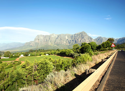 The Old Franschhoek Mountain Pass Scenic rural road along a hillside with green fields, a wooden guardrail, and distant mountains under a clear blue sky.