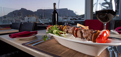 Three Days in Cape Town day 1 Outdoor dining table with a skewered grilled kebab platter, greens, and peppers, plus a bottle and glass of red wine; harbor and mountains in the background.