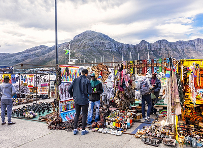 Three Days in Cape Town day 3 Outdoor market stall along a waterfront, with colorful textiles, hats, scarves, and souvenirs as shoppers browse against a mountainous backdrop.