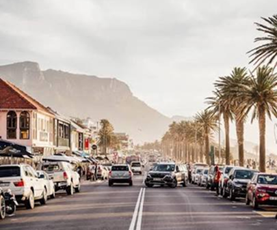 Victoria Road – Camps Bay to Hout Bay Busy coastal street with parked cars, palm trees, and a mountain backdrop in the distance.