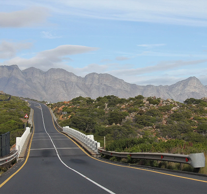 Why Scenic Drives Are Better at a Relaxed Pace Winding two-lane road with guardrails curves through scrubby hills toward distant rocky mountains under a blue sky.